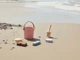 En lyserød spand og legetøjssandforme er anbragt på en sandstrand nær kysten med blide bølger og spredte småsten i baggrunden.