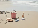 En lyserød spand, en legetøjsskovl og farverige sandforme står på sandstranden nær kysten med bølger i baggrunden.
