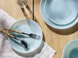 Lyseblå tallerkener med gaffel og ske på den ene, sat på et træbord med en stribet dug, et glas vand og en skål, der er delvist synlig.