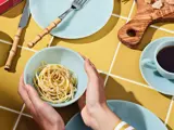 Hænderne holder en skål spaghetti over et gult flisebord med tallerkener, bestik, en kop kaffe og et træbræt med ost.