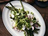 En tallerken med frisk salat med asparges, radiser i skiver, grønne ærter og bladgrønt på et hvidt ovalt fad med blå kant.
