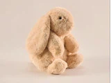 En blød, beige kanin i plys med hængeører og sorte øjne sidder oprejst på en almindelig, lys baggrund.