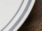 Nærbillede af en hvid tallerken med to grå striber nær kanten, placeret på en brun træoverflade.