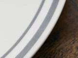 Nærbillede af en hvid tallerken med to grå striber nær kanten, placeret på en brun træoverflade.