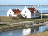 Et hvidt hus med trappegavle og rødt tag står ved vandet med et dansk flag på en stang og et spejlbillede i en nærliggende bæk.