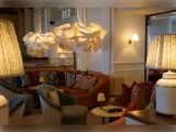 Hyggelig lounge med plyssofaer, mønstrede puder, skulpturelle pendler, bordlamper og et skakbræt på et lille bord, som skaber en varm og indbydende atmosfære.