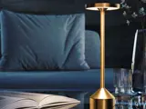 En moderne bordlampe i guld er tændt på et bord ved siden af en åben bog, et glas og en vase med en blå sofa og pude i baggrunden.
