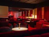 En svagt oplyst lounge med røde lænestole, runde borde og små bordlamper, røde gardiner på væggene og en bar i baggrunden.