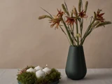 En mørk vase med spidse røde blomster står ved siden af en krans af mos, hvor der står fire hvide stearinlys – hvoraf det ene er tændt – på et lyst bord foran en beige baggrund.