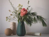 En grøn vase med forskellige vilde blomster og fyrretræsgrene står ved siden af et stykke sejlgarn, en beige kugle og en mønstret rulle, alt sammen på en grå overflade foran en lys væg.
