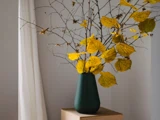 En mørkegrøn vase med grene med gule efterårsblade står på et bord af lyst træ foran en ensfarvet væg og et hvidt gardin.