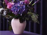 En lyserød vase rummer en livlig buket af lilla, blå og rosa blomster med mørkegrønne blade på en dyb lilla baggrund.