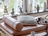 En moderne daybed i læder med et cylindrisk bolster og en struktureret pude står ved et vindue, omgivet af bøger, dekorative vaser og et hyggeligt tæppe.