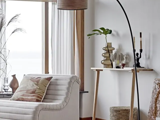 En moderne stue med hvid sofa, mønstret pude, gulvlampe, konsolbord med dekoration, flettet kurv og stort vindue med gennemsigtige gardiner.