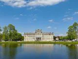En storslået historisk bygning med udsmykket arkitektur står bag en dam, omgivet af grønne træer under en blå himmel med spredte skyer.
