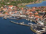 Luftfoto af en kystby med bygninger med røde tage, en marina med både og omkringliggende grønne områder og vand.