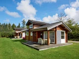 Tre små træhytter med verandaer ligger på en grøn græsplæne, omgivet af træer, under en blå himmel med spredte skyer.