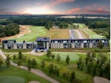 Et moderne hotel med store vinduer ligger ved siden af en golfbane, omgivet af grønne fairways, træer og en dramatisk solnedgangshimmel.