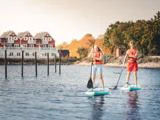 To personer i redningsveste på paddleboard på roligt vand nær en kystlinje med røde og hvide huse og træer i baggrunden.