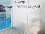 Glasdør med dansk tekst Lysterapi - Kig ind og se lyset. Bag døren er der en indendørs swimmingpool med blåt vand og et farverigt vægmaleri.