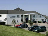 En hvid toetagers bygning med navnet "Løgstør Parkhotel" med en parkeringsplads foran og flere parkerede biler, omgivet af grønt græs og små træer.