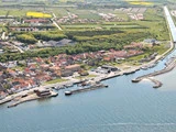 Luftfoto af en kystby med en marina, dokker og både, omgivet af huse, grønne områder og landbrugsjord i baggrunden.
