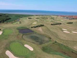 Luftfoto af en kystnær golfbane med grønne fairways, sandbunkers, små damme og havet i baggrunden under en klar blå himmel.