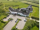 Luftfoto af en stor, hvid, L-formet bygning med en parkeringsplads omgivet af grønne marker og træer. Flere biler er parkeret uden for hovedindgangen.