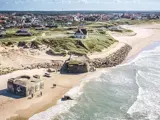 Luftfoto af en sandstrand med bunkere fra 2. verdenskrig, græsklædte klitter, spredte huse og en kystby i baggrunden. Bølger møder blidt kystlinjen.