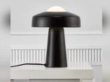 En moderne sort bordlampe med en bred, kuppelformet skærm og en cylindrisk fod står på et hvidt bord mod en lysegrå væg.