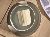 En beige kop placeret i en lysegrå skål, begge anbragt på en større grå og beige tallerken oven på en træoverflade.