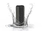 En sort bærbar Bluetooth-højttaler sprøjtes med vand, hvilket fremhæver dens vandtætte funktion, mod en hvid baggrund.