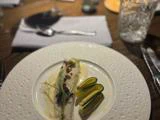 En anrettet gourmetret med en cremet sauce, mikrogrønt og tyndtskårne gule og grønne grøntsager, anrettet på et træbord med bestik og et glas ved siden af.