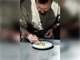 En kok i en beige uniform bruger en pincet til omhyggeligt at anrette maden på en hvid tallerken, mens han koncentrerer sig fuldt ud om anretningen.