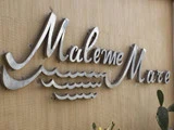 Sølvfarvet metalskilt med teksten Maleme Mare og bølgemotiver på en beigefarvet væg.