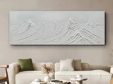 Et hvidt maleri med struktur, der forestiller bjergtoppe på et rektangulært lærred, hænger over en moderne beige sofa med neutrale og grønne puder og minimalistisk indretning.