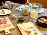 Et træbord dækket med ost, salami, brød, marmelade, en dessert, juice og skåle med tilbehør, arrangeret til et måltid.