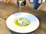 En brun sauce hældes fra en lille kande over et fad med fisk, fennikel i skiver og grøn sauce på en hvid tallerken på et træbord.