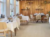Moderne restaurantinteriør med trævægge, lyse træstole, hvide duge, pænt dækkede borde og dekorative planter i nærheden af store vinduer.