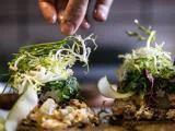 En hånd drysser frisk grønt på smørrebrød med forskellige grøntsager og pålæg og viser en detaljeret præsentation.