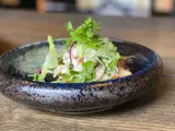 På et træbord står en rustik keramikskål fyldt med en frisk salat, toppet med bladgrønt og krydderurter.