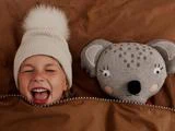 Et grinende barn med en hvid strikhue ligger i sengen ved siden af en grå, udstoppet koala, begge gemt under et brunt tæppe.