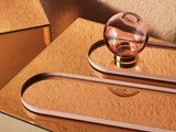 En lyserød glaskugle med guldbund sidder på en skinnende kobberoverflade og reflekterer lyset. Der er også buede lyserøde metalstrimler på overfladen, som skaber en moderne, abstrakt komposition.