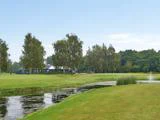 En græsklædt golfbane med en lille dam og et springvand i forgrunden, træer og en klubhusbygning i baggrunden under en klar himmel.