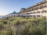 Et stort strandhotel med balkoner og solstuer i glas, omgivet af højt græs og sandklitter under en klar himmel.