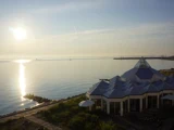 En moderne bygning med glastag nær roligt vand ved solopgang eller solnedgang, med en skarp sol, der reflekterer i havet, og en klar himmel over.