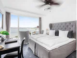 Lyst hotelværelse med en stor seng, skrivebord, stole og en balkon med udsigt over vandet og byens skyline. Store vinduer lukker naturligt lys ind.