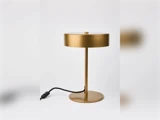 En moderne bordlampe i guld med en rund, flad lampeskærm og fod, der står på en hvid baggrund med en sort strømledning fastgjort.