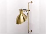 En vægmonteret lampe i messing med en buet arm og en kuppelformet skærm, fastgjort til en hvid væg, med en synlig sort ledning.