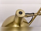 Nærbillede af en messingfarvet skrivebordslampe med en sort tænd/sluk-knap på toppen og en justerbar arm i baggrunden.